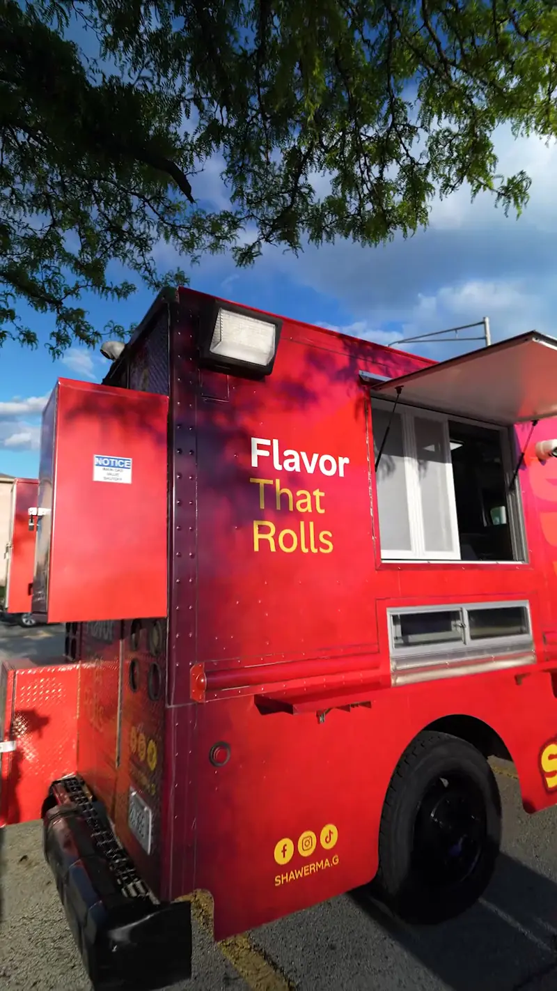 The Shawerma G truck parked under a tree, its "Flavor That Rolls" panel and service window facing the lot